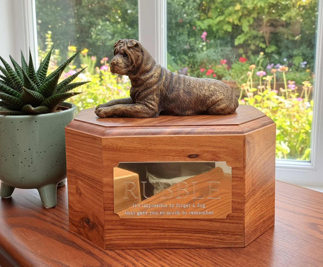 Wooden urn with a bronze dog figurine on top, placed on a wooden surface with a plant and blanket in the background.