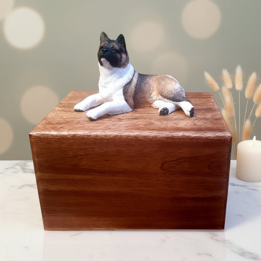 A wooden urn with a sculpture of an Akita on top, hand painted and in a lying pose.