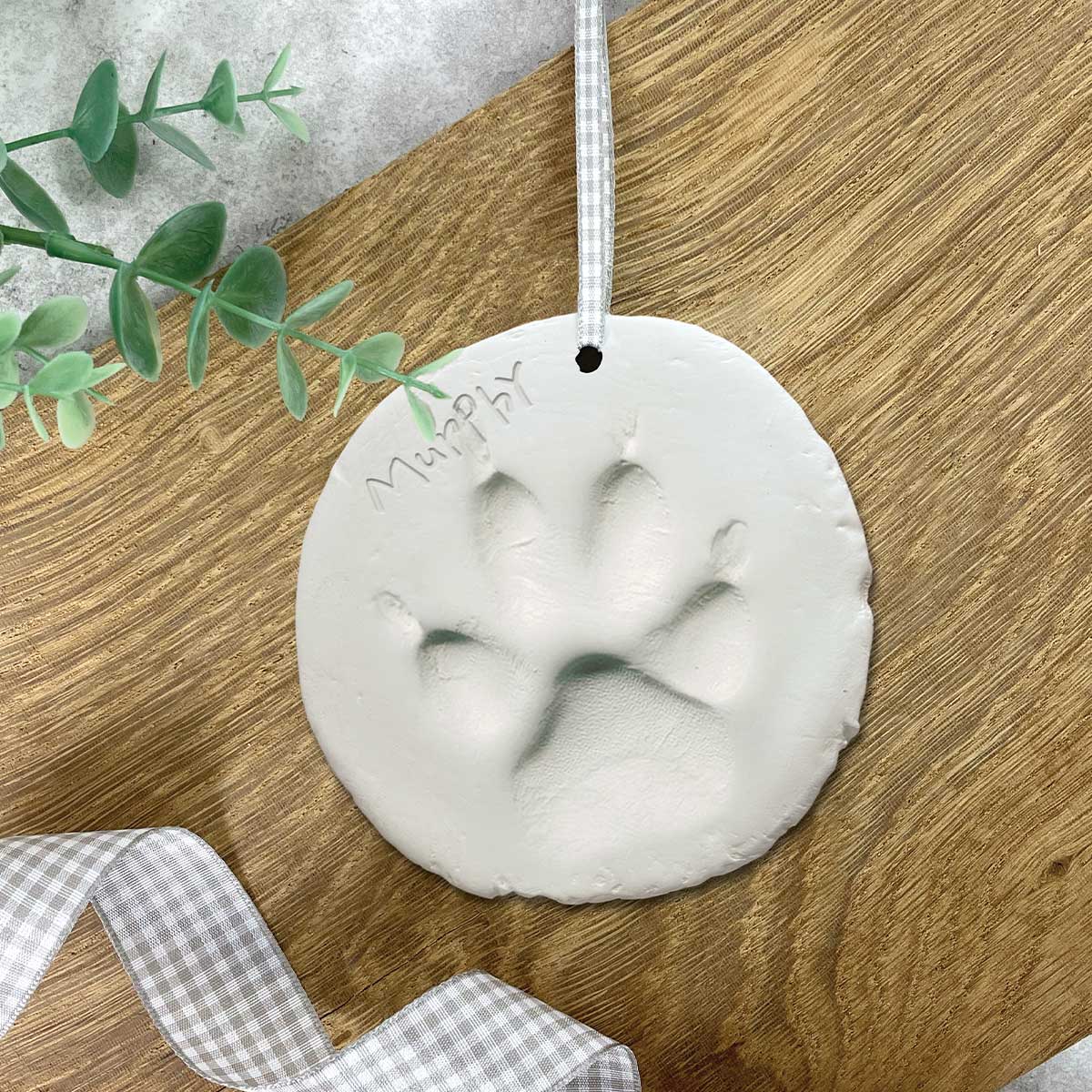 Round clay ornament with paw print on a wooden surface with greenery and ribbon.