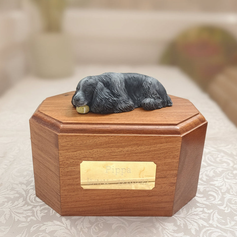 Wooden urn with a small figurine on top, placed on a bed.