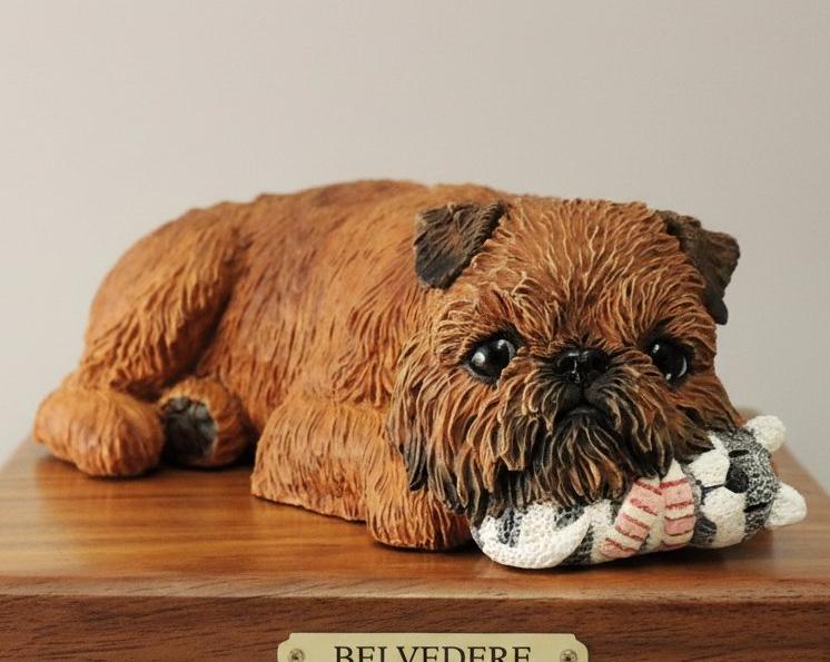 A hand-painted wooden urn sculpture in the shape of a Griffon Bruxellois dog lying down, with a cream and grey scarf in its mouth.